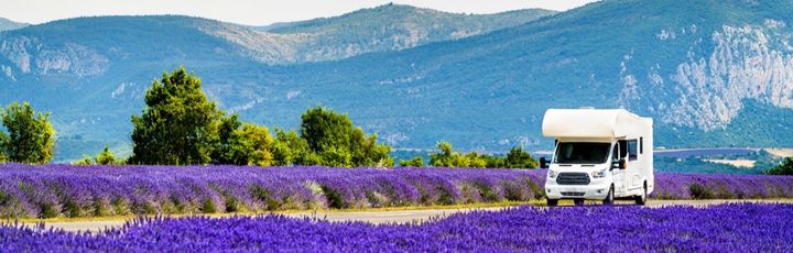 Ein weißer Campervan parkt am Rand eines blühenden Lavendelfeldes mit lila Blüten und lila Bergen im Hintergrund.
