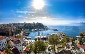Panoramablick auf den historischen Hafen von Antalya, Türkei, mit traditionellen Segelschiffen, der Altstadt und dem glitzernden Mittelmeer unter strahlender Sonne.