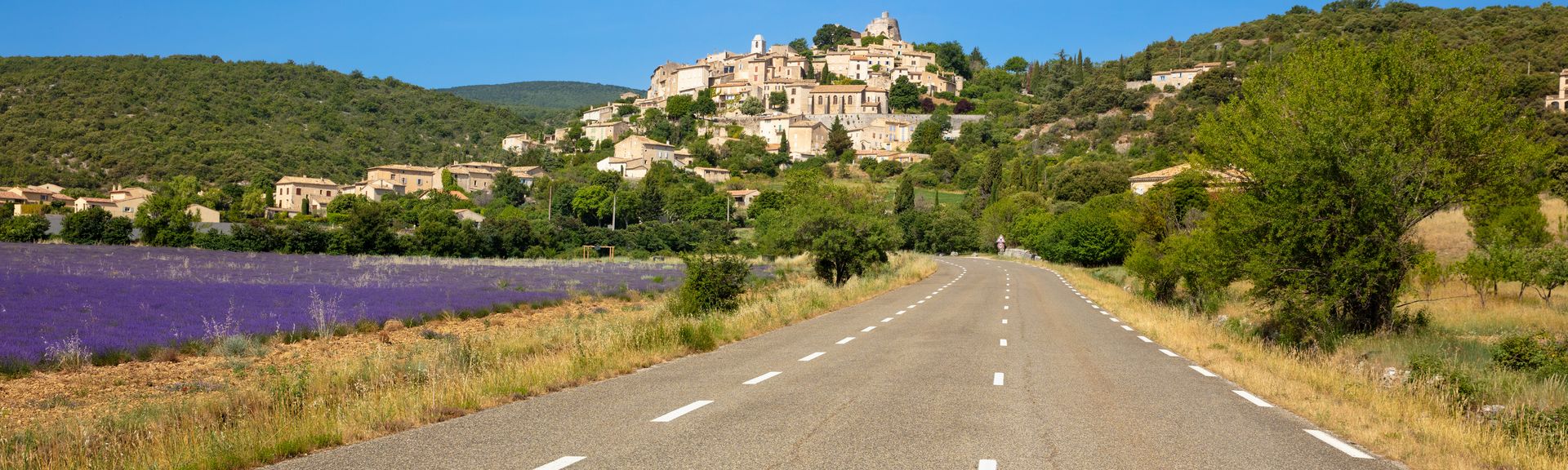 Eine idyllische provenzalische Landschaft im Luberon, mit einem charmanten Dorf, das auf einem Hügel thront, umgeben von grünen Feldern und sanften Hügeln unter einem klaren blauen Himmel.