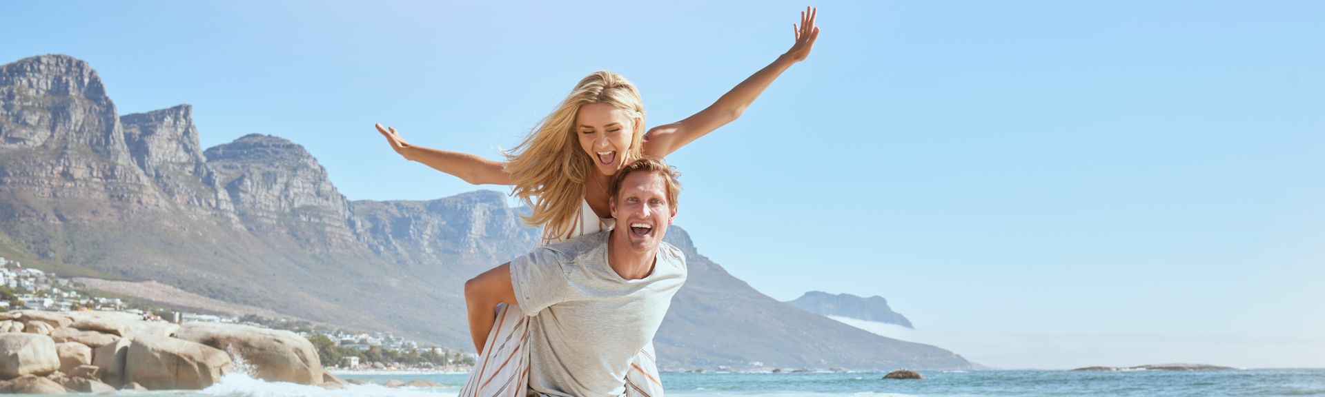 Paar lauft fröhlich am Strand, Frau sitzt auf Rücken vom Mann und streckt Hande in die Höhe