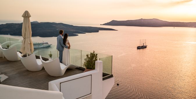 Paar steht auf dem Balkon eines griechischen Luxushotels und blickt im Sonnenuntergang auf das Meer 