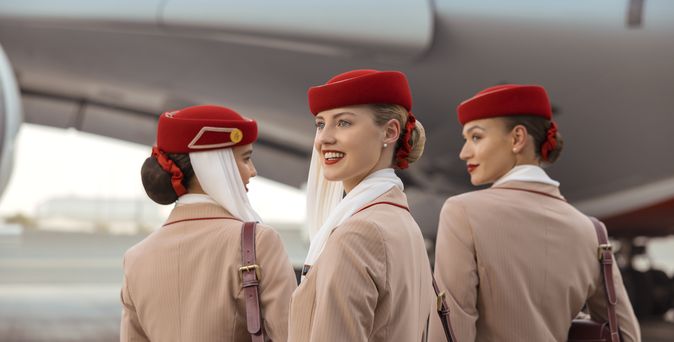 Drei Emirates-Flugbegleiterinnen in Uniform mit roten Hüten stehen nebeneinander auf dem Rollfeld vor einem Flugzeug.