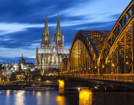 Der Kölner Dom und die Hohenzollernbrücke bei Nacht, beleuchtet, spiegelt sich im Rhein.