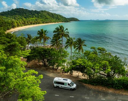 Camper steht an einer Straße mit Palmen und Sträuchern, dahinter befindet sich ein langer Strand mit türkisblauem Meer in Australien