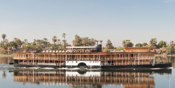 Ein historisches Dampfschiff, die SS Sudan, fährt auf dem Nil, umgeben von üppiger grüner Vegetation und dem ruhigen Wasser.