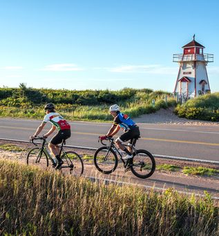 2 Radfahrer, die eine Strasse entlang einer Küstenlandschaft fahren , mit Leuchtturm im Hintergrund