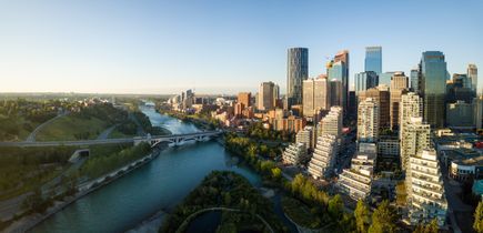 Die Stadt Calgary. Viele Hochhäuser, teilweise direkt am Fluss mit kurzer Brücke, blauer Himmel, Sonnenuntergang. In der Ferne im Nebel kann man die Berge, die Rocky Mountains, erkennen.