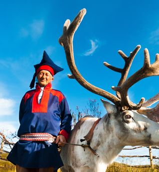 Eine Person in traditioneller samischer Kleidung steht neben einem Rentier in einer verschneiten Landschaft.