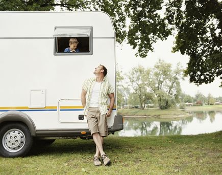 Ein Vater und sein Sohn sitzen auf Stühlen vor einem Campervan, der Sohn liest ein Buch.