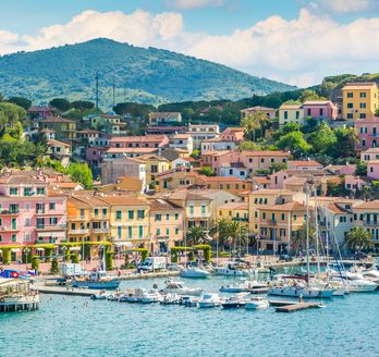 Blick auf Porto Azzuro vom Wasser aus. In der Front viele kleine Boote, dahinter viele schnuckelige bunte Häuser. Dahinter wiederum ein Berg und blauer Himmel.