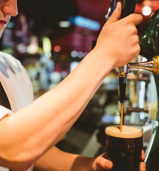 Ein Barkeeper in einem weißen Hemd und einer dunklen Krawatte, der mit beiden Händen ein dunkles Bier aus einem Zapfhahn in ein Pint-Glas zapft. Der Arm des Barkeepers ist im Vordergrund, und im Hintergrund ist eine Barumgebung unscharf zu erkennen.