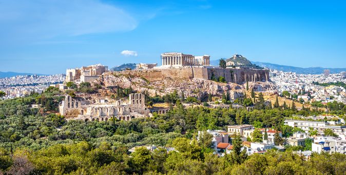 Blick über Athen. Oben auf dem Berg ist die Akropolis zu sehen. Unterhalb viele weiße Häuser, umgeben von Bäumen. Himmel ist strahlend blau.