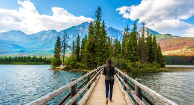 Eine Wanderung geht über einen Holzsteg, welcher an den Seiten über Baumstämme als Abgrenzung verfügt und über einen türkisblauen Gletschersee führt. Blick auf die wundervolle Berg-und Fauna-Landschaft.