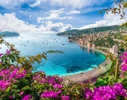 Blick über die Bucht, Häuser im Hintergrund, Küstenstraße zu sehen, Blumen im Vordergrund. Klares Wasser, blauer Himmel