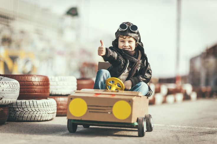 Junge trägt Fliegerbrille und Lederjacke und fährt in einem selbstgebauten Pappkarton-Auto mit erhobenem Daumen auf einer Rennstrecke.