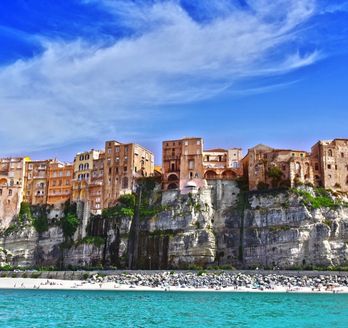 Die Stadt Tropea in Italien thront auf einer steilen Klippe über dem türkisfarbenen Meer, mit einem historischen Gebäude auf einem Felsen im Vordergrund.