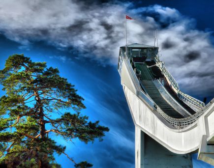 Skisprungschanze bei blauem Himmel. Ein hoch gewachsener Nadelbaum ist ebenfalls zu sehen.