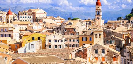 Blick auf die Altstadt von Korfu-Stadt (Kerkyra) in Griechenland mit ihren charakteristischen Terrakotta-Dächern, engen Gassen und dem markanten Glockenturm von Saint Spyridon vor einem Hintergrund aus Bergen und blauem Himmel mit weißen Wolken.