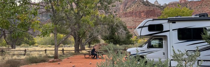Ein weißes Wohnmobil ist auf einem sandigen Campingplatz unter Laubbäumen geparkt. Im Vordergrund befindet sich ein Picknicktisch mit einer Person, die daran sitzt. Im Hintergrund erheben sich rötliche Felsformationen unter einem leicht bewölkten Himmel.