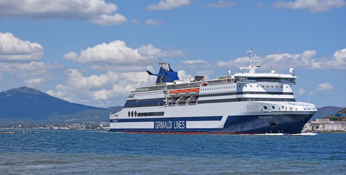 Ein großes weißes und blaues Fährschiff mit der Aufschrift "Grimaldi Lines" auf der Seite fährt auf dem Meer mit Bergen im Hintergrund.
