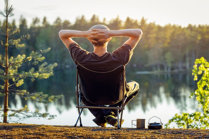 Eine Person sitzt entspannt in einem Holzstuhl am Wasser.