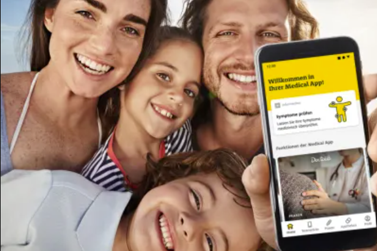 Glückliche Familie macht ein Selfie, im Vordergrund Smartphone mit geöffneter ADAC Medical App, Symbol für digitale Sicherheit.