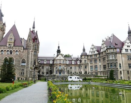 Das Schloss Moszna in Polen, ein historisches Schloss und Palast mit romantischer Märchen- und eklektischer Architektur, umgeben von einem Wasserspiegel und Gärten unter einem bewölkten Himmel