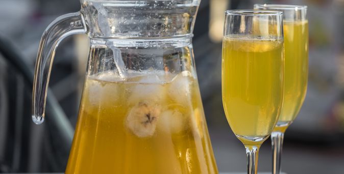 ein helles Getränk in der Karaffe, daneben zwei gefüllte Sektgläser, auf einem Tisch stehend. 