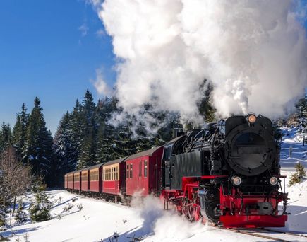 Eine Dampflokomotive der Harzer Schmalspurbahnen fährt durch eine verschneite Winterlandschaft, umgeben von dichten Wäldern und Bergen.