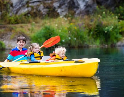 Zwei Kinder paddeln in einem gelben Kanu auf einem ruhigen Fluss, umgeben von grüner Natur.