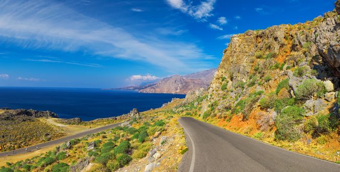 Eine Küstenstraße welche Richtung tiefblauem Meer führt. Links und Rechts trockene Landschaft mit ein wenig grünen Büschen. Am Himmel sind kleine Wölkchen zu sehen, ansonsten strahlend blau.
