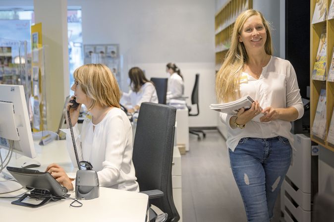 Büro mit zwei Frauen. Eine Frau sitzt am Tisch und telefoniert. Die andere Frau hat etwas in der Hand und läuft lächelnd im Gang entlang