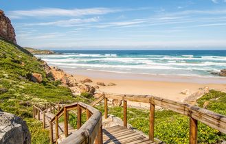 Eine weite Küstenlandschaft des Robberg Nature Reserve in Südafrika, mit zerklüfteten Klippen, die ins blaue Meer abfallen, und einer Sandbank, die sich in die Ferne erstreckt.