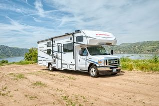  Ein weißes El Monte RV Wohnmobil der Klasse C ist auf einem unbefestigten Platz in der Nähe eines Gewässers und hügeliger Landschaft unter einem leicht bewölkten Himmel geparkt.