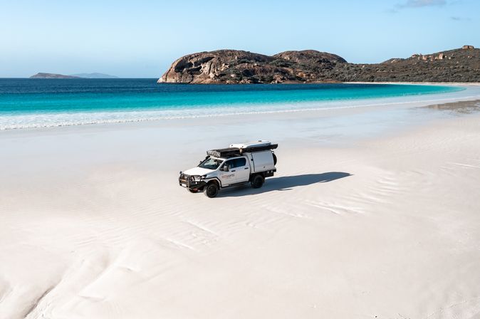 Red Sands weißer Camper steht an einem weißen Strand mit türkisem Meerwasser und im Hintergrund Horizont und Felsen.