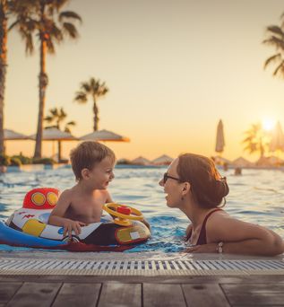 Mutter und Kind mit Schwimmreif im Pool. Im Hintergrund Palmen und Sonnenuntergang.