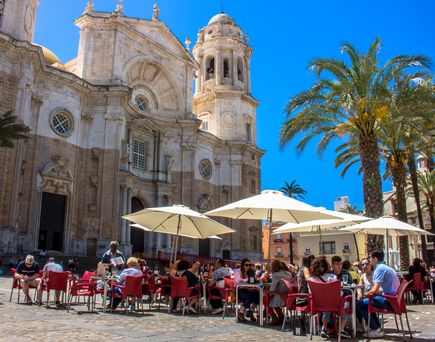 Marktplatz Cadiz. Davor einige Stühle, Tische und Sonnenschirme. Die Gäste sitzen dort und verzehren etwas. Im Hintergrund eine Kapelle, Palmen und blauer Himmel