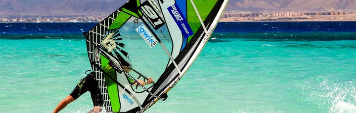 Ein Windsurfer auf dem türkisfarbenen Wasser, der ein grünes Segel mit einer roten und weißen Grafik auf einem sonnigen Tag benutzt.