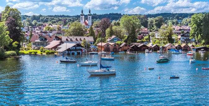 Starnberger See in Bayern mit Booten und im Hintergrund ein Dorf mit Kirche und Bäumen.