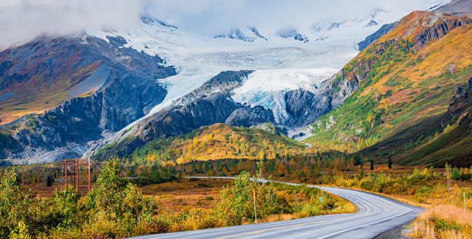 Eine kurvenreiche Straße führt durch eine herbstliche Landschaft mit gelb und orange gefärbten Bäumen und Sträuchern, die zu einem großen Gletscher mit schneebedeckten Bergen im Hintergrund führt. Der Himmel ist teilweise bewölkt.