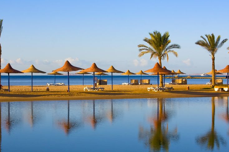 Ein Sandstrand mit Reihen von Strohsonnenschirmen und Liegestühlen, die sich an einem sonnigen Tag an einem Hotelresort in Hurghada, Ägypten, widerspiegeln.