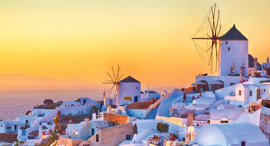 Weiß getünchte Häuser und Windmühlen des Ortes Oia auf Santorini leuchten im goldenen Licht der Abendsonne, mit Blick auf das Ägäische Meer.