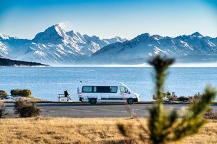 Ein weißer Mercedes-Benz Sprinter Maxi Taxi steht auf einer gepflasterten Straße in einer städtischen Umgebung, umgeben von Gebäuden und einem weiten, blauen Himmel.