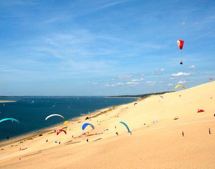 Ein ausgedehnter Sandstrand mit einer Person, die sich entspannt, und einem bunten Drachenflieger, der unter einem klaren blauen Himmel schwebt. Das Meer ist hellblau und ruhig.