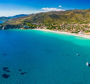Blick über türkisblaues Meer, Strand und etwas Wald im Hintergrund. Blauer Himmel.