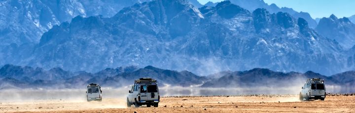 Mehrere weiße Geländewagen fahren im Konvoi durch die staubige Wüste vor einer eindrucksvollen, bläulichen Bergkette.