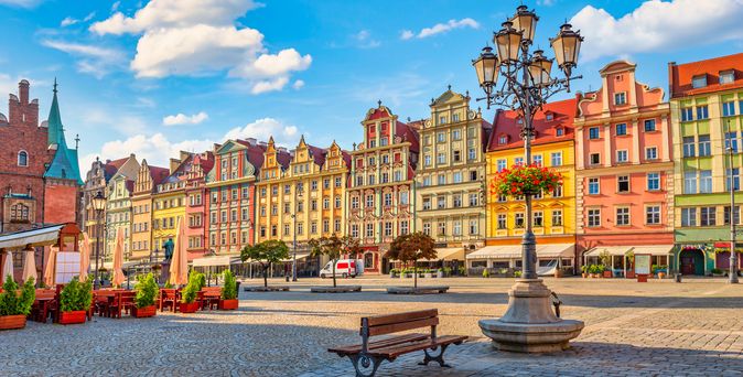Zu sehen ist die bunte Altstadt von Breslau mit seinem schönem Marktplatz und einem Brunnen und einer Bank im Vordergrund.
