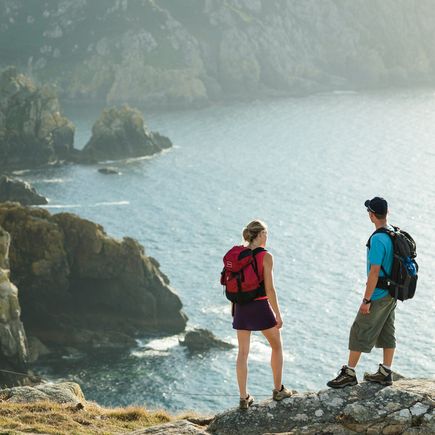 Zwei Personen stehen an der Küste und blicken auf das weite, ruhige Meer. Die Landschaft ist bewölkt, und die Farben sind gedämpft, was eine nachdenkliche Stimmung erzeugt.