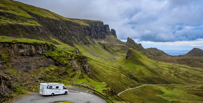 Camper fährt um eine Kurve in einem grünen Gelände in Schottland mit felsigen Bergen und weit im Hintergrund das Meer.