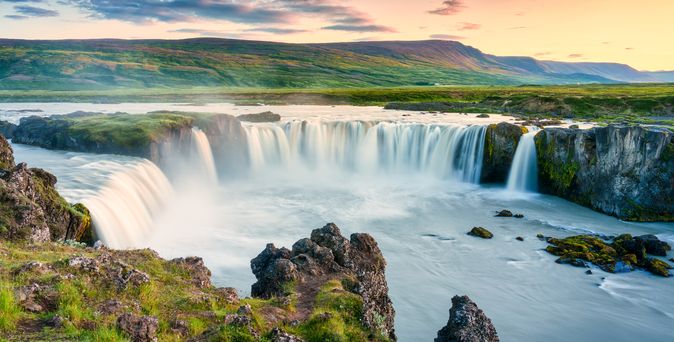 Ein Wasserfall in einer grünen Landschaft mit Bergen im Hintergrund.